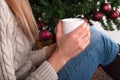 Young Girl in sweater holding cup of coffee in hand at legs with warmers and Christmas tree in the background