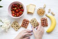 Young girl takes vegan toast with fruits, seeds, peanut butter over white wooden background, top view. Healthy breakfast with ingr