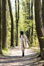 Young girl with trenchcoat and hat walks away down the road in the spring sunny forest, back view