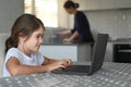 Young girl using a laptop at home while her mother cleaning the kitchen Royalty Free Stock Photo