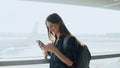 Young girl using smartphone near airport window. Happy European woman with backpack uses mobile app in terminal. 4K. Royalty Free Stock Photo