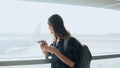 Young girl using smartphone near airport window. Happy European woman with backpack uses mobile app in terminal. 4K. Royalty Free Stock Photo