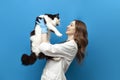 young girl veterinarian in uniform holds black and white cat on blue isolated background, doctor in medical coat