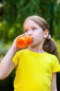 Young girl in a vibrant yellow t-shirt outdoors enjoying a refreshing drink from a clear bottle filled with orange carrot juice. Royalty Free Stock Photo