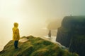 Young girl wearing raincoat standing on the edge of a cliff with huge waves rolling ashore. Rough foggy Irish weather. Beautiful