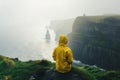 Young girl wearing raincoat standing on the edge of a cliff with huge waves rolling ashore. Rough foggy Irish weather. Beautiful