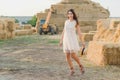 Young girl wears summer white dress near hay bale in field. Beautiful girl on farm land. Wheat yellow golden harvest in autumn Royalty Free Stock Photo