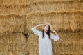 Young girl wears summer white dress near hay bale in field. Beautiful girl on farm land. Wheat yellow golden harvest in autumn Royalty Free Stock Photo