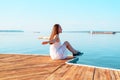 Young girl in white dress sitting on a wooden pier with open hands looking into the distance of the sea, freedom, clean air, dream Royalty Free Stock Photo