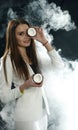 Young girl in a white jacket holds a coconut near her eye and smiles on a black background, covered with smoke vapor