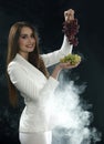 A young girl in a white jacket holds a grapes in her hands and smiles on a black background, covered with smoke vapor Royalty Free Stock Photo