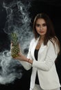 A young girl in a white jacket holds a pineapple in her hands and smiles on a black background, covered with smoke vapor