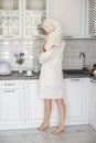 A young girl in a white robe and with a towel on her head stands in a white kitchen