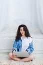 Young girl in white t-shirt and denim shirt, sits on the floor near the bed and uses a laptop. Royalty Free Stock Photo