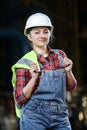 Young girl in a work dress and white hard hat in a factory. Woman in a work uniform holding reflective vest in a storage.