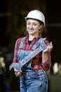 Young girl in a work dress and white hard hat holding big wrench and hummer in a factory. Woman in a work uniform.