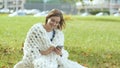 A young girl wrapped in a merino plaid typing on the phone a message sitting on the grass in a city park. Royalty Free Stock Photo