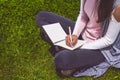 Young girl writes with pen in notebook sitting on green grass in park on meadow Royalty Free Stock Photo