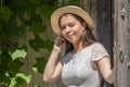 A young girl of 17-20 years old in a white dress and a straw hat smiles and opens a wooden door, leaving a rural house. Royalty Free Stock Photo