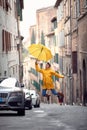 A young girl with a yellow raincoat and umbrella is jumping on the street while enjoying a walk through the city on a rainy day.