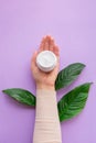 Young girls hand is nearby jar with white soft hand and body cream on purple background with large green leaves.Concept of eco cos Royalty Free Stock Photo