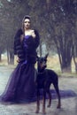 Glam lady in luxurious sequin violet evening gown and fur coat standing on the alley in the park with her Doberman pinscher dog