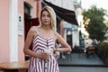 Young glamorous woman blonde in a vintage pink striped dress with a milkshake in hands stand on the street near open cafe.