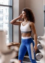 Young good fit curly girl dressed in sports clothes is standing next to the sports equipment in the modern gym by the