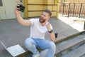 Young good looking bearded guy is sitting outdoors on stairs in front of his house with laptop, making selfie and showing rock Royalty Free Stock Photo