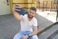 Young good looking bearded guy is sitting outdoors on stairs in front of his house making selfie and smiling. Wearing white shirt Royalty Free Stock Photo