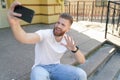 Young good looking bearded guy is sitting outdoors on stairs in front of his house making selfie and smiling. Wearing white shirt Royalty Free Stock Photo