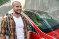 Young good looking bold bearded guy standing outdoors near his red car smiling. wearing yellow shirt