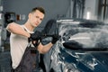 Young good-looking man worker washing luxury dark blue car on a car wash, looking at camera and holding high pressure