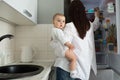 Young good-looking mother with dark hair holding newborn son on hands and looking for baby food in opened refrigerator