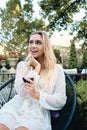 Young gorgeous blond woman in white dress dreamily looking away with glass of red wine in garden of restaurant Royalty Free Stock Photo