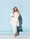 Young gorgeous platinum blonde woman in white pants, blouse, coat and high-heeled shoes walking towards camera