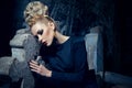 Young gothic woman in black dress with makeup and hairstyle and crown, poses near a cross, in a medieval castle