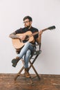 Young guitarist hipster at home with guitar on the chair full-body portrait