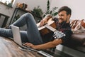 Young guitarist hipster at home sitting on the floor holding guitar earphones music browsing laptop Royalty Free Stock Photo