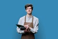 Young guy in apron making notes on clipboard, on blue studio background Royalty Free Stock Photo