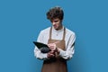 Young guy in apron making notes on clipboard, on blue studio background Royalty Free Stock Photo