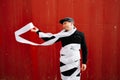 A young guy in black sports clothes and a baseball cap on the street, standing near a red wall wrapped in toilet paper ribbons