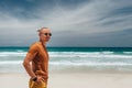 A young guy in a brown T-shirt and shorts stands on the sandy seashore in sunglasses. Clear summer day, blue sky with white clouds Royalty Free Stock Photo