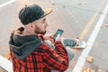 A young guy in a cap, with a beard, holds a smartphone and looks to the right. In the background is a road and a skateboard. Back