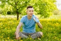 Young guy dressed in a blue t-shirt, drinks water from a bottle, sits on a green grass and yellow dandelion background Royalty Free Stock Photo