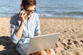 Young guy with glasses, working on his laptop and talking on the phone on the beach, working on vacation, suitable for advertising Royalty Free Stock Photo