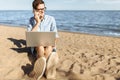 Young guy with glasses, working on his laptop and talking on the phone on the beach, working on vacation, suitable for advertising Royalty Free Stock Photo