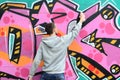 A young guy in a gray hoodie paints graffiti in pink and green c