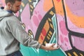 A young guy in a gray hoodie paints graffiti in pink and green c