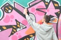 A young guy in a gray hoodie paints graffiti in pink and green c